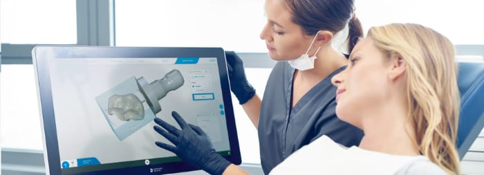 A dental patient and tech are looking at a CEREC machine computer monitor that displays a 3D image of the patient's dental crown restoration, which will be manufactured in-house in about one hour.